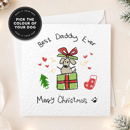 A white Christmas greeting card with a hand-drawn illustration of a dog sitting on a green gift box with a red bow, surrounded by small red hearts, a green tree, a red stocking, and the text 'Best Daddy Ever' and 'Merry Christmas'. A black circular badge in the top left says 'Pick the colour of your dog' and 'Make it your own'.
